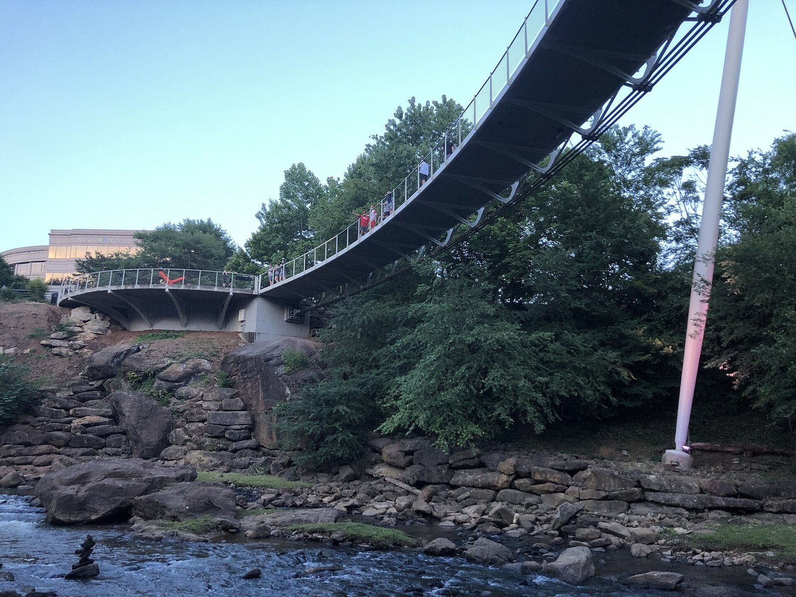 The curved Liberty Bridge seen from below, hanging over rocks and the Reedy River, with a tall white support mast