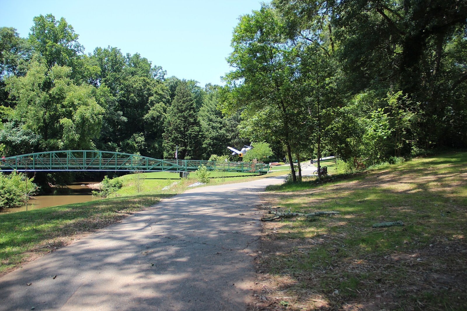 A paved trail curving through green trees toward a green metal footbridge in Greenville