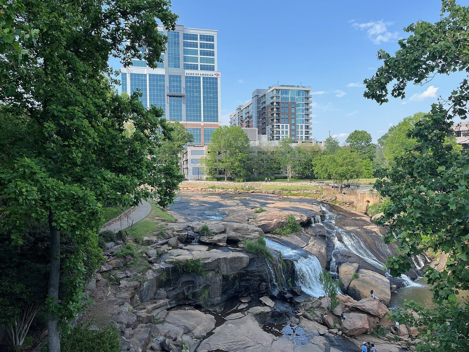 Reedy River Falls running over wide brown rock ledges in Falls Park, with downtown Greenville buildings above the trees