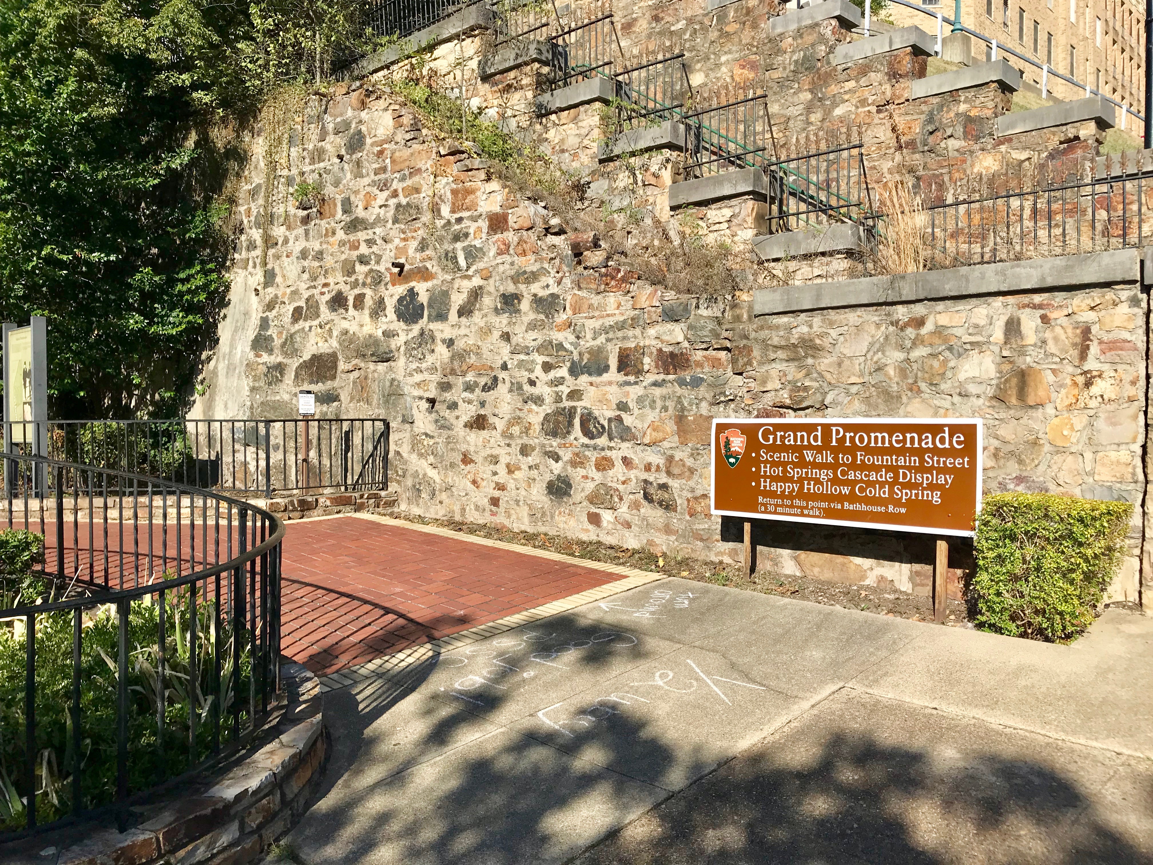 The brick Grand Promenade trailhead in Hot Springs National Park, with a stone retaining wall and a National Park Service sign listing free features