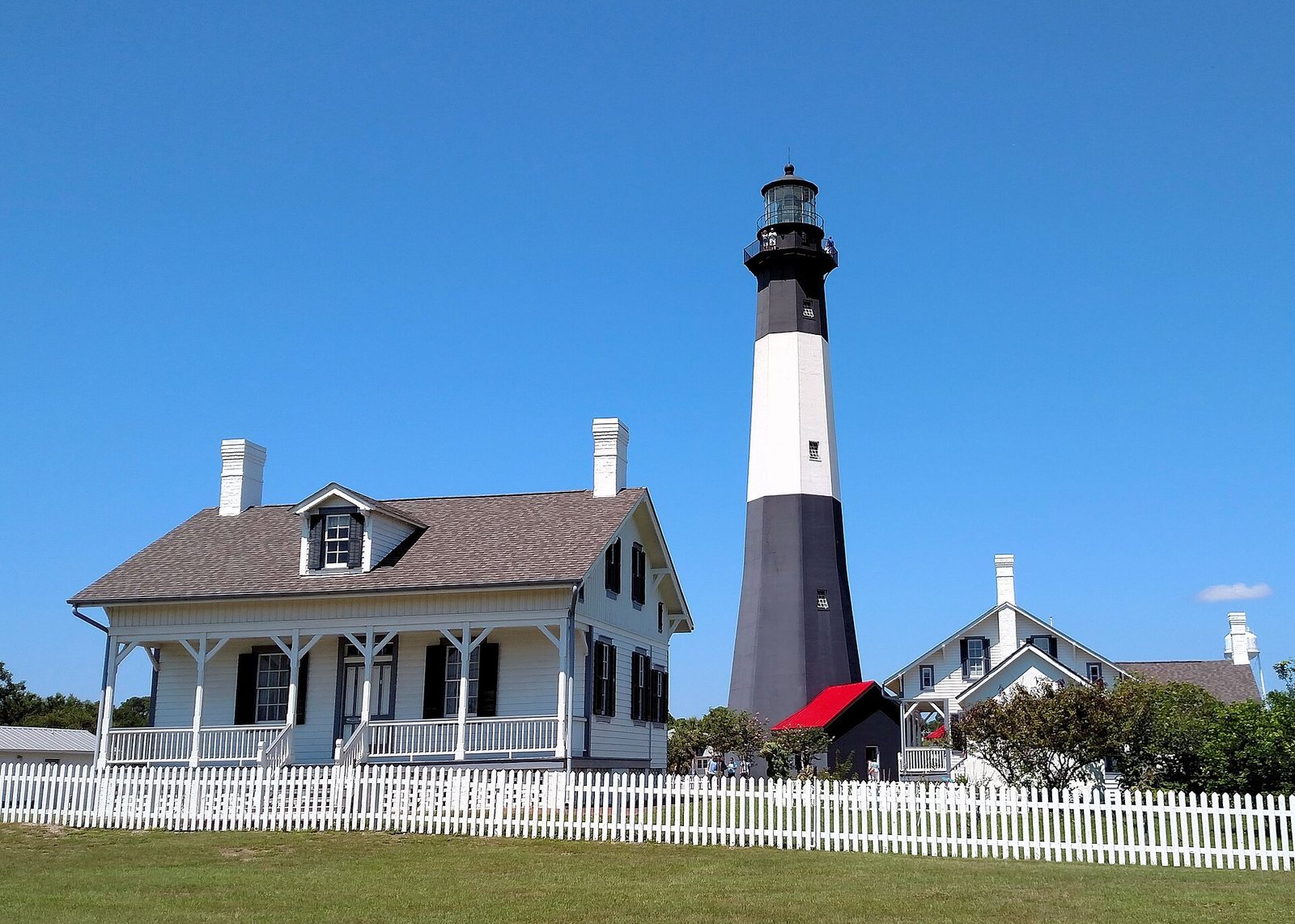 The black and white Tybee Island lighthouse beside the white lightkeeper's house and a picket fence under a clear blue sky